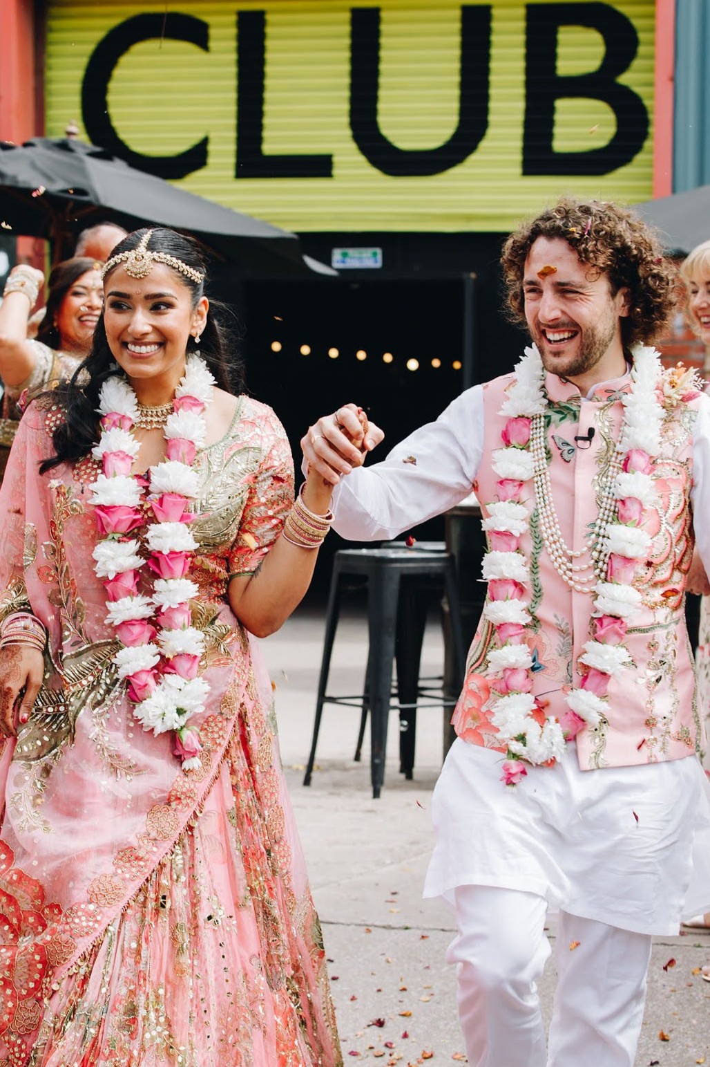 Will and Jasmine on their wedding day, hand in hand and laughing, both wearing floral garlands.